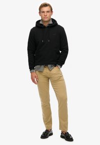 Sudadera negra con cordones, bolsillo frontal, superpuesta sobre una camisa de cuadros. Pantalones beige y mocasines negros completan el atuendo.