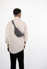 Borsa a tracolla grigia in tessuto testurizzato, indossata sulla schiena. Camicia oversize beige con maniche larghe e pantaloni neri. Design pulito e minimalista.