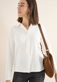 Weißes Langarm-Hemd mit Knopfleiste und Kragen, kombiniert mit einer Schultertasche aus braunem Wildleder. Einfaches Design mit glatter Textur.