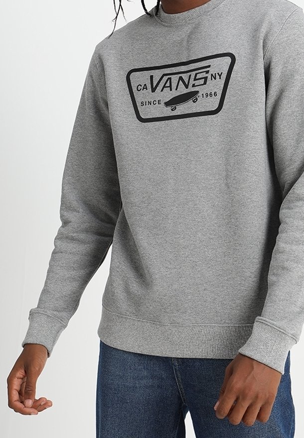 Felpa grigia Vans con logo nero e grafica di skateboard indossata da una persona in jeans blu, mostrata dalla spalla ai fianchi.