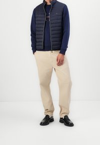 Homme portant un gilet matelassé bleu marine sur un sweat-shirt bleu marine, un pantalon beige et des mocassins en cuir noir, debout devant un fond blanc.