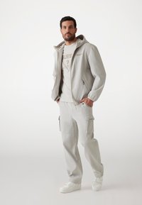 Ljusgrå zip-jacka med ribbad textur, tillsammans med matchande ribbade byxor. Klädseln inkluderar en vit logot-shirt och vita sneakers.