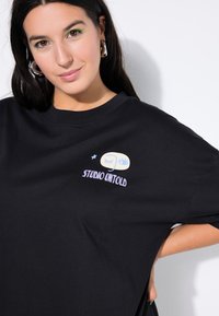 Camiseta de algodón negra con un corte relajado, que presenta un diseño gráfico de ojos y el texto "STUDIO UNTOLD" en púrpura.