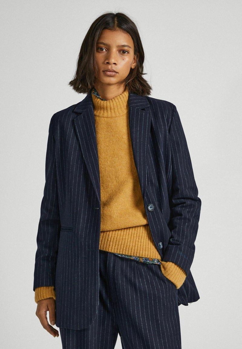 Blazer a rayas marinas con dos botones, combinado con un suéter de canalé mostaza. Corte a medida, con acentos decorativos en el dobladillo interior.