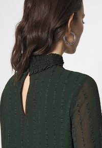 Robe vert foncé avec un col montant froncé, des manches transparentes et un détail en trou de serrure dans le dos, présentant des rayures verticales texturées.