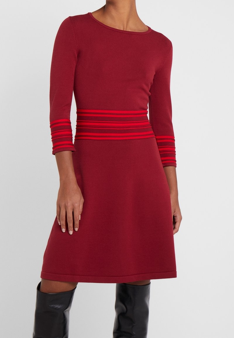 Femme portant une robe évasée bordeaux avec des accents rayés rouges à la taille et aux manches, associée à des bottes noires jusqu'au genou.