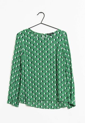 Groene blouse met lange mouwen, een ronde halslijn en een zwart-wit patroon van halve cirkels, hangend aan een zwarte kledinghanger tegen een witte achtergrond.