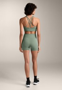 Zelená športová podprsenka s prekríženými ramienkami na chrbte a zodpovedajúcimi krátkymi nohavicami s vysokým pásom. Materiál vyzerá mäkký a elastický, vhodný na športové oblečenie.