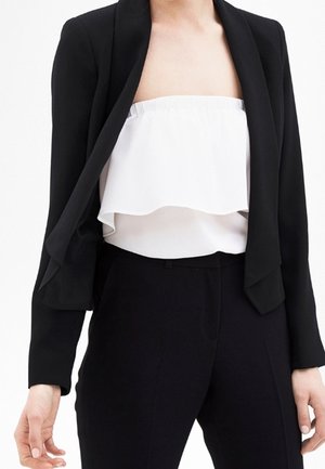 Kobieta w czarnym blezerze na białej, bezramieniowej, falbaniastej bluzce i czarnych spodniach z wysokim stanem, stojąca na białym tle.