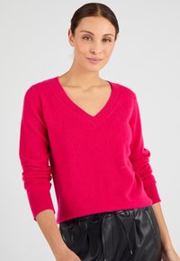 Pull en V fuchsia à manches longues, fabriqué à partir d'un matériau doux. Les poignets côtelés ajoutent de la texture. Associé à un pantalon noir plissé avec cordon de serrage.