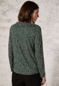 Langsleeve top in donkergroen met witte en teal zigzagpatronen. Zacht materiaal met een ronde halslijn en een relaxed fit, stijlvol aan de achterkant.