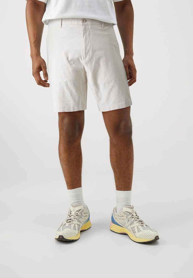 Homme portant un short blanc, des chaussettes blanches et des baskets athlétiques blanches, jaunes et bleues, debout sur un fond clair uni.
