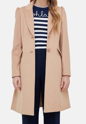 Vrouw draagt een beige wollen jas met één knoop over een marineblauw-wit gestreepte trui met de tekst "oh la", gecombineerd met marineblauwe broek.