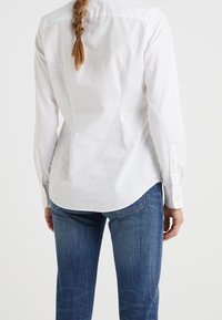 Vit bomullsskjorta med långa ärmar, knäppning vid ärmsluten och en rundad nederkant. Bärs med blå denimjeans, vilket visar upp en enkel och ren design.