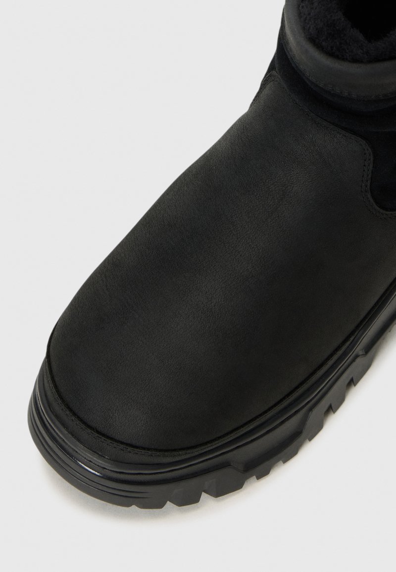 Schwarzer Knöchelstiefel mit elegantem Design, strukturiertem Obermaterial und einer klobigen Sohle mit Profilmuster, vor einem neutralen Hintergrund.