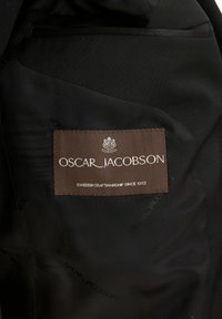Interno di un blazer nero con un'etichetta marrone che riporta "OSCAR JACOBSON" e "SWEDISH CRAFTSMANSHIP SINCE 1903" in testo bianco. Motivo a spina di pesce.