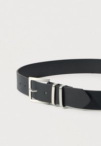 Μαύρη ζώνη από συνθετικό δέρμα με ασημί ορθογώνια αγκράφα, δύο καρφιά και πολλαπλές τρύπες για ρύθμιση μεγέθους. Λείος υφής, μίνιμαλ σχεδιασμός.