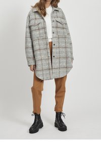 Grå og brun ternet oversized skjortejakke med to brystlommer, rund hac, båret over en hvid top og parret med brune bukser og sorte støvler.