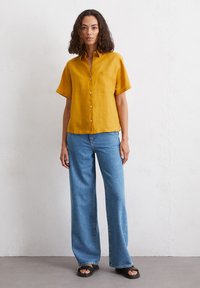 Mosterdkleurig overhemd met korte mouwen en een borstzak, gecombineerd met lichtblauwe wijde jeans en zwarte sandalen.