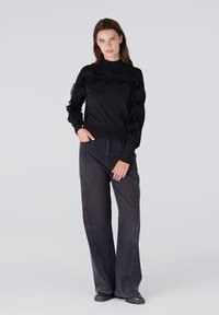 Zwarte sweater met ruches op de schouders, lange mouwen en een hoge halslijn, gecombineerd met een donkerdenim jeans met hoge taille.