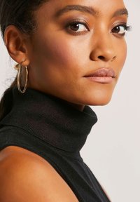 Schwarzer Rollkragenpullover mit gerippter Textur und hohem Kragen. Eine goldene Creole betont das Ohr in einem Nahaufnahme-Porträt.