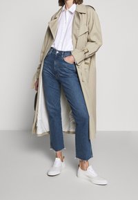 Trench beige sobre una camisa blanca de botón, combinado con unos vaqueros de tiro alto azules y zapatillas blancas. Los vaqueros tienen dobladillos deshilachados.