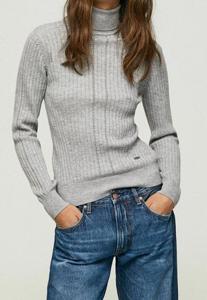 Femme portant un pull à col roulé côtelé gris clair et un jean en denim bleu, les mains dans les poches, vue du cou jusqu’aux hanches.
