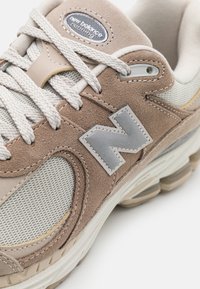 Chaussure de sport beige et grise avec des matériaux en suède et en mesh, dessus texturé, fermeture à lacets et un accent logo argenté proéminent.