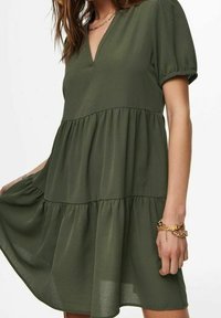 Femme portant une robe à volants olive à manches courtes avec un décolleté en V, accessoirisée d'un collier chaîne doré et de bracelets au poignet droit.