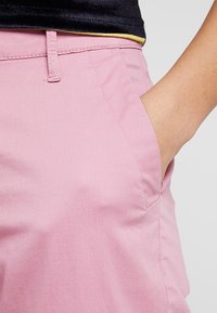 Ljusrosa bomullsbukser med en sidoficka, jämn textur och skräddarsydd passform, med synlig midjeband och sömdetaljer.