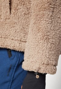 Beiger Fleece-Pullover mit weicher, plüschiger Textur und rundem Halsausschnitt. Bündchenärmel mit verstellbaren Knöpfen. Kombination mit blauen Hosen.