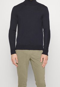 Marinblå stickad polotröja med långa ärmar och ribbade muddarna, kombinerad med khakifärgade byxor. Tröjan har en figurnära design och en slät yta.