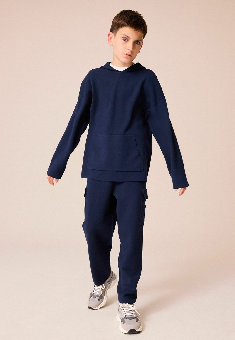 Dunkelblaues Sweatshirt mit Kapuze und Fronttasche, kombiniert mit dunkelblauen Cargo-Hosen. Graue und schwarze Sneakers mit weißen Schnürsenkeln. Schlicher Hintergrund.