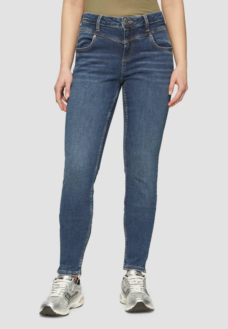 Jean skinny en denim dans un lavage bleu foncé, avec cinq poches, passants de ceinture et couture contrastante. Associé à des baskets de sport argentées.