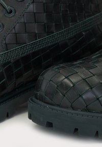 Black braided leather boots with a textured finish, featuring a chunky rubber sole and dark green accents. Laces and metal eyelets visible.