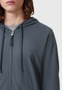 Felpa grigia con zip e cappuccio regolabile, texture morbida e un piccolo disegno ricamato sul lato sinistro del petto. Il tessuto sembra leggero.