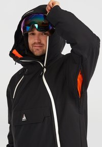 Giacca nera con colletto alto, cerniera bianca e un'accente arancione all'interno del cappuccio. Presenta una tasca frontale e una texture del tessuto adatta per l'abbigliamento outdoor.