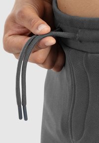 Pantalons de survêtement gris avec une taille ajustable par cordon, présentant des liens en tissu tressé et une ceinture texturée. Gros plan sur une main tenant le cordon.