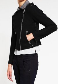 Svart kort jacka med silverfärgade dragkedjor och grå krage, med en texturerad tyg och sidofickor. Bärs över en grå tröja.