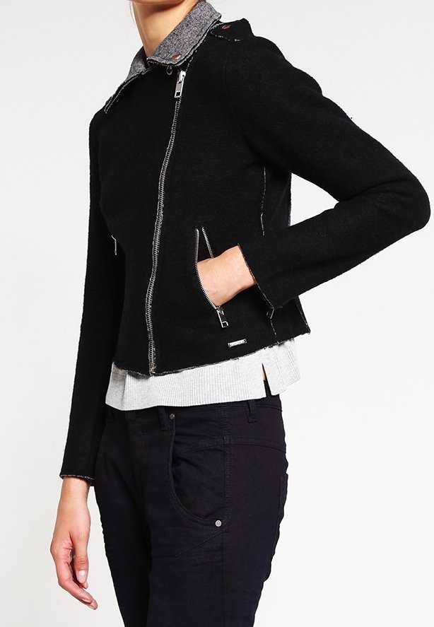 Svart kort jacka med silverfärgade dragkedjor och grå krage, med en texturerad tyg och sidofickor. Bärs över en grå tröja.