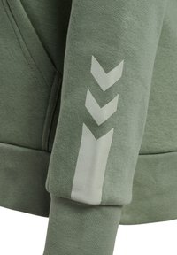 Manche de sweat-shirt verte avec trois chevrons blancs pointant vers le bas près du poignet et une couture de poche visible.