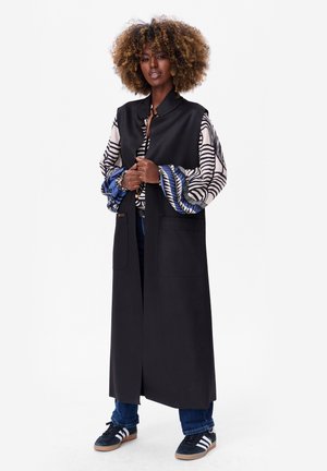 Long gilet noir avec deux poches avant, porté par-dessus une chemise à motifs avec des rayures bleues et blanches, associé à un jean bleu et des baskets noires.