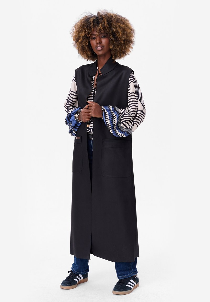 Long gilet noir avec deux poches avant, porté par-dessus une chemise à motifs avec des rayures bleues et blanches, associé à un jean bleu et des baskets noires.