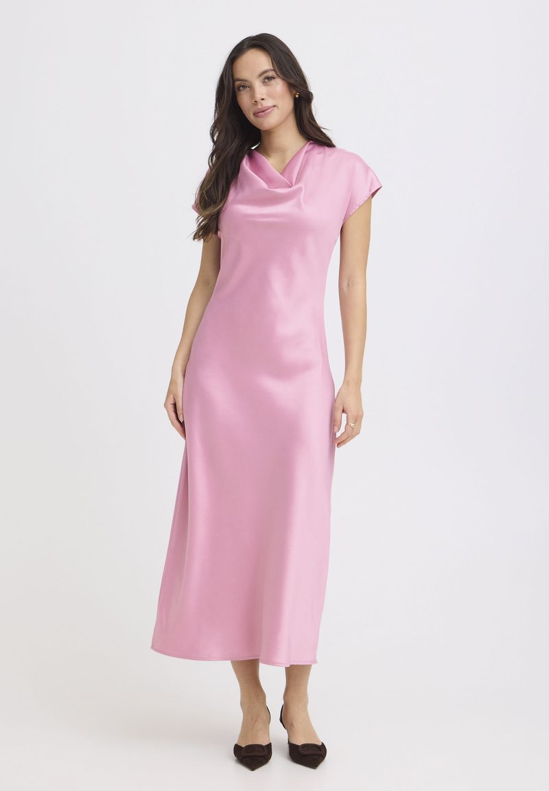 Kobieta z długimi ciemnymi włosami, ubrana w różową satynową sukienkę midi bez rękawów i czarne szpilki z ostrym czubkiem, stojąca na białym tle.