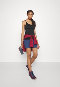 Svart linne, blå shorts, röd hoodie knuten i midjan, håller i en grå vattenflaska. Mörka sneakers med turkosa snören. Rent bakgrund.