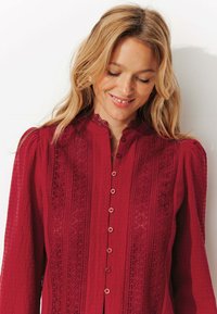 Chemise rouge à boutons, ornée de détails en dentelle complexes, avec un tissu texturé et un col mandarin agrémenté de manches froncées.