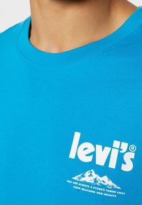 Gros plan d'un t-shirt bleu Levi's avec logo blanc et graphisme montagne accompagné du texte « Vous êtes toujours à un jet de pierre d'atteindre de nouveaux sommets. »