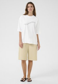 Witte oversized T-shirt met een zwart spikkeldesign, gecombineerd met beige knielange shorts en zwarte sandalen. Glad katoenen textuur.