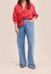 Donna che indossa una camicia rossa larga con bottoni, infilata parzialmente nei pantaloni blu a vita alta e a gamba larga, con cintura marrone e scarpe aperte nere.