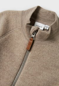 Beige zip-up jacka i mjukt material med hög krage, ribbade detaljer och en läderdragkedja med silverfärgad hårdvara.
